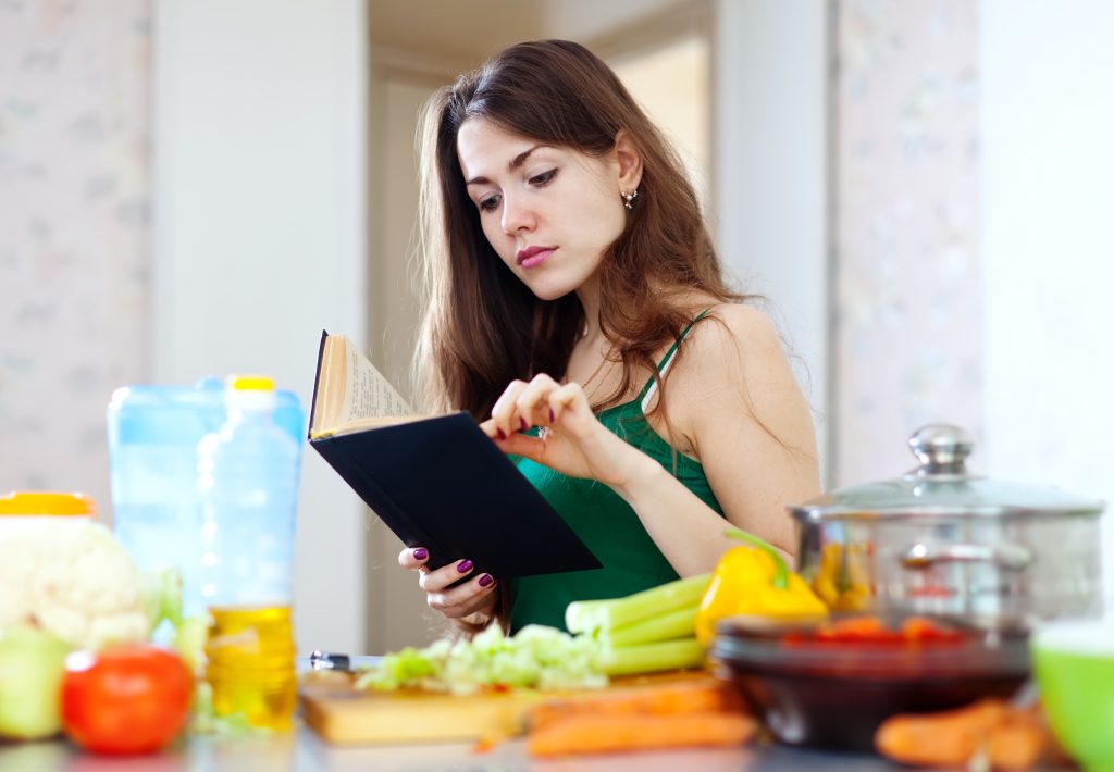 Young woman reading a cookbook in the kitchen while preparing fresh vegetables and Fruits