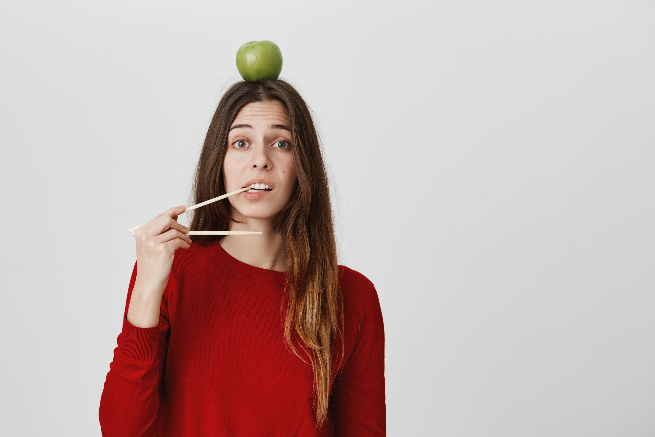 Woman in red sweater holding chopsticks near her mouth with a green apple balanced on her head.