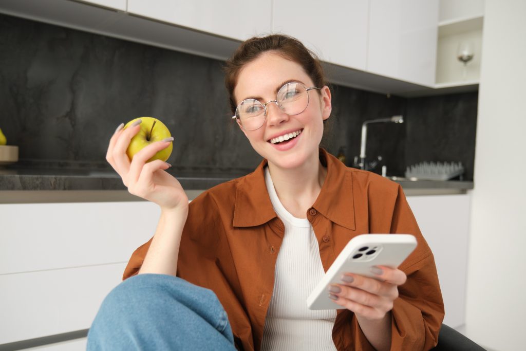 a happy, beautiful young woman in glasses, sitting with mobile phone, eating apple, laughing and smiling, and counting calories in apple
