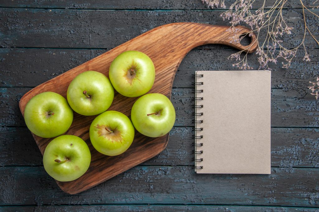Green apples on a kitchen board with a notebook on a dark surface, showing a healthy lifestyle